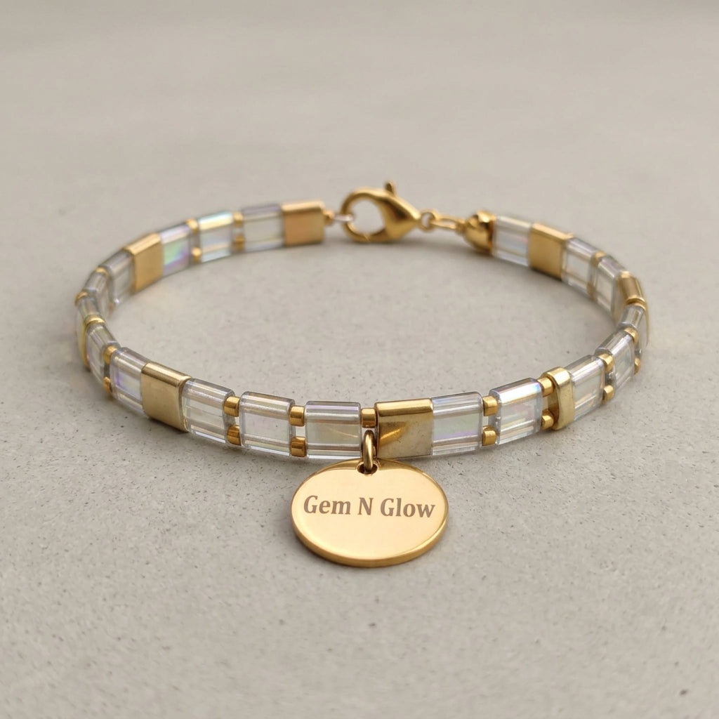 Clear iridescent and gold-tone Miyuki Tila bracelet with a round Gem N Glow charm on pale fabric.