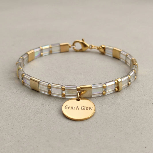 Clear iridescent and gold-tone Miyuki Tila bracelet with a round Gem N Glow charm on pale fabric.