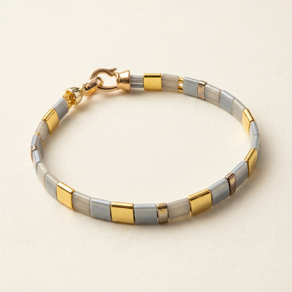 Grey and gold Miyuki Tila and Delica bracelet with a gold-tone clasp on a white surface.